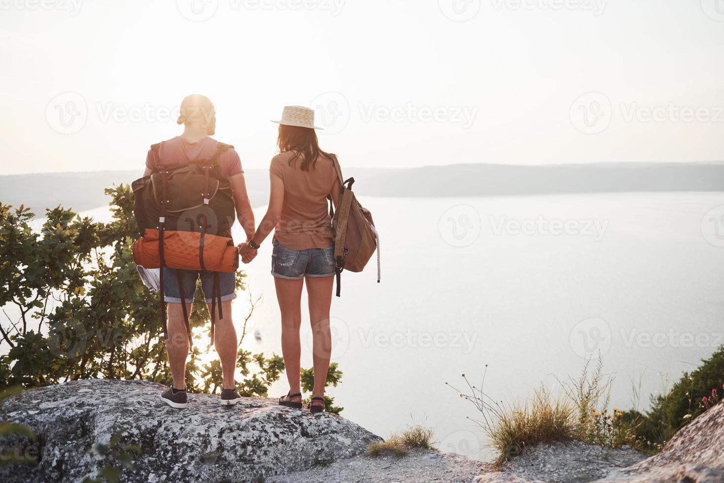 två turist man och kvinna med ryggsäckar står på toppen av klippan och njuter av soluppgången. resande berg och kust, frihet och aktivt livsstilskoncept foto