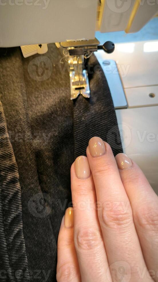 bearbeta av sömnad i en sömnad maskin sy kvinnas händer manikyr svart textil- denim jeans tillverkning stänga upp sömmerska i verkstad tyg kläder framställning design hobby handgjort arbetssätt handarbete foto