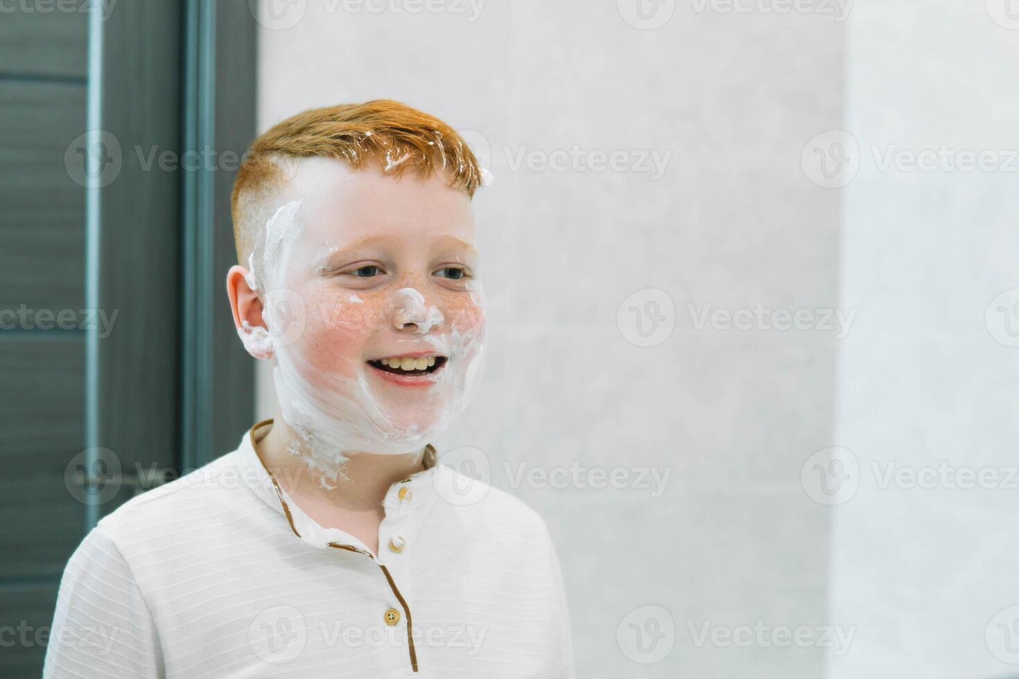 rolig pojke i de bad utstryker hans ansikte med rakning grädde, de barn imiterar ett vuxen pappa rakar sig. de pojke insmord hans kinder med rakning skum foto