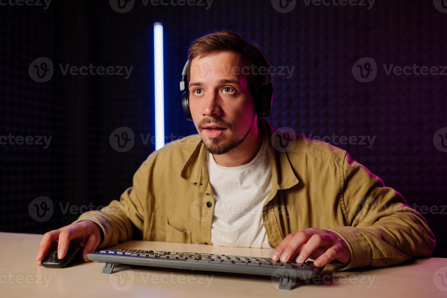 stilig man i skjorta kläder och headsetet Sammanträde på en tabell i en rum med neon lampor och spelar spel på de dator med en leende på hans ansikte ser på de skärm. foto