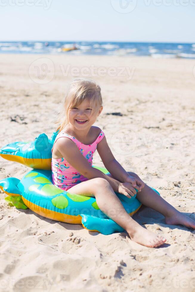 Lycklig flicka av europeisk utseende ålder av 5 Sammanträde och skrattande på ett uppblåsbar krokodil leksak på de strand sommar solig dag.familj sommar yrke begrepp. vertikal Foto. foto