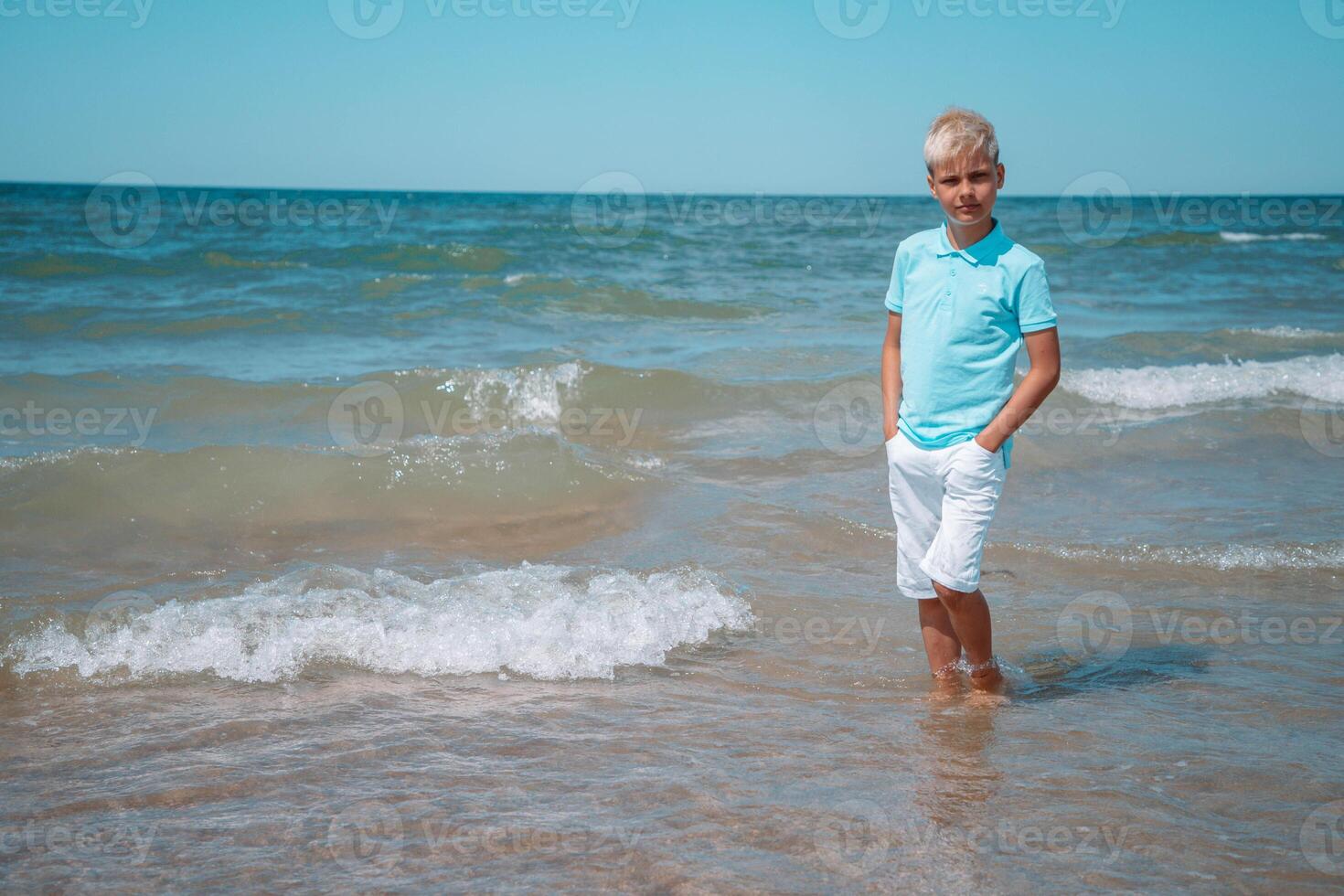 stilig tonåring pojke av europeisk utseende med blond hår i vit shorts, och en blå t-shirt står i de hav i de vatten, och utseende till de kamera. sommar semester koncept.sommar resa , familj högtider, resa koncept.kopia Plats. foto