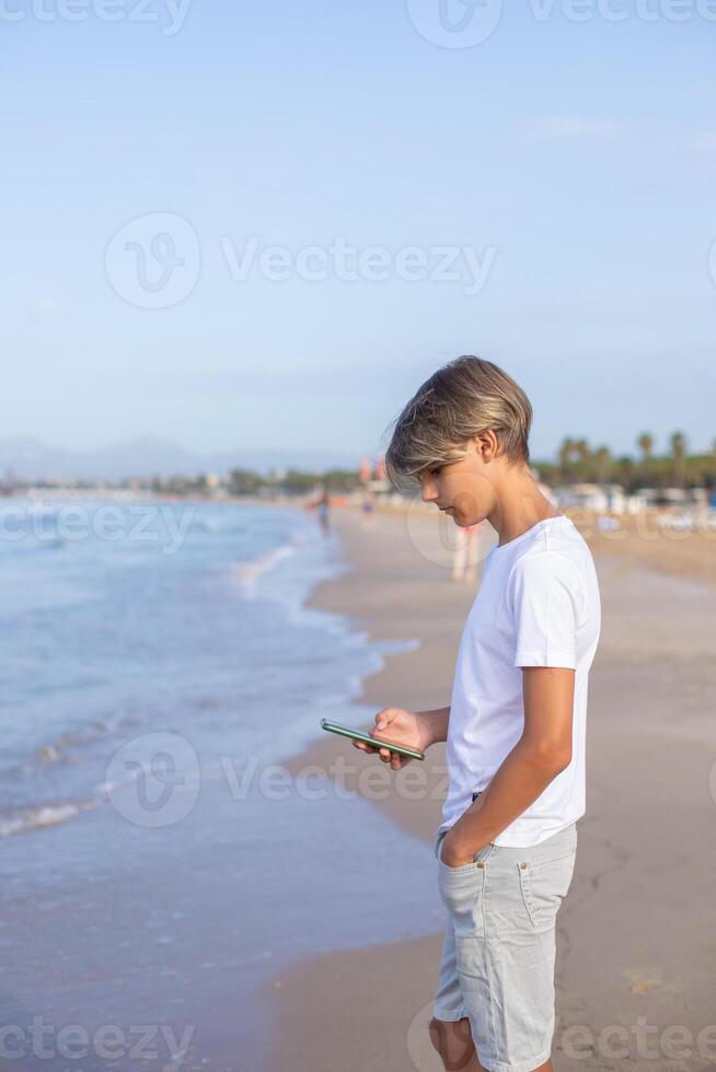 stilig tonåring pojke i vit t-shirt använder sig av smartphone på de strand på sommar högtider, resa eller resa i Spanien .resa,semester,semester, frihet koncept.sida se vertikal . foto