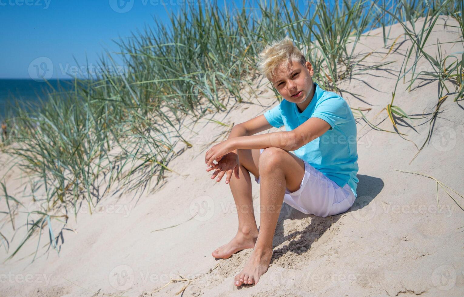 stilig tonåring pojke av europeisk utseende med blond hår i vit shorts, och en blå t-shirt sitter på en strand, och utseende till de kamera. sommar semester koncept.sommar resa koncept.kopia Plats. foto