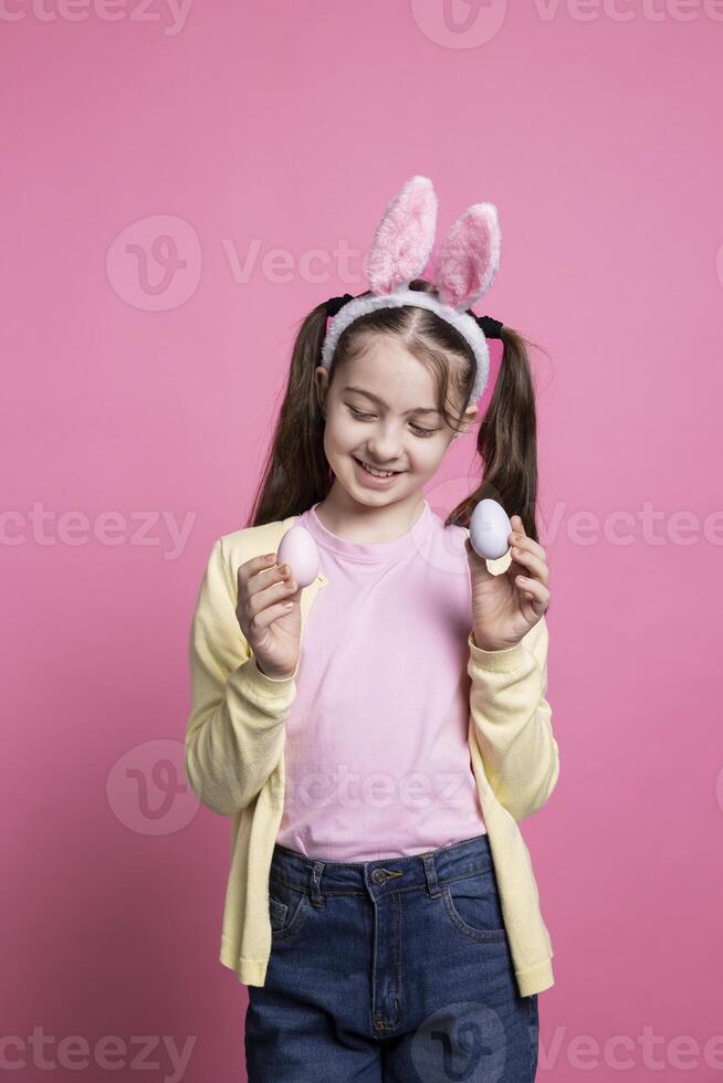 små barn Framställ med förtroende i främre av kamera, som visar henne målad färgrik ägg för påsk firande. ung ljuv unge med kanin öron leende i studio, presenter handgjort dekorationer. foto
