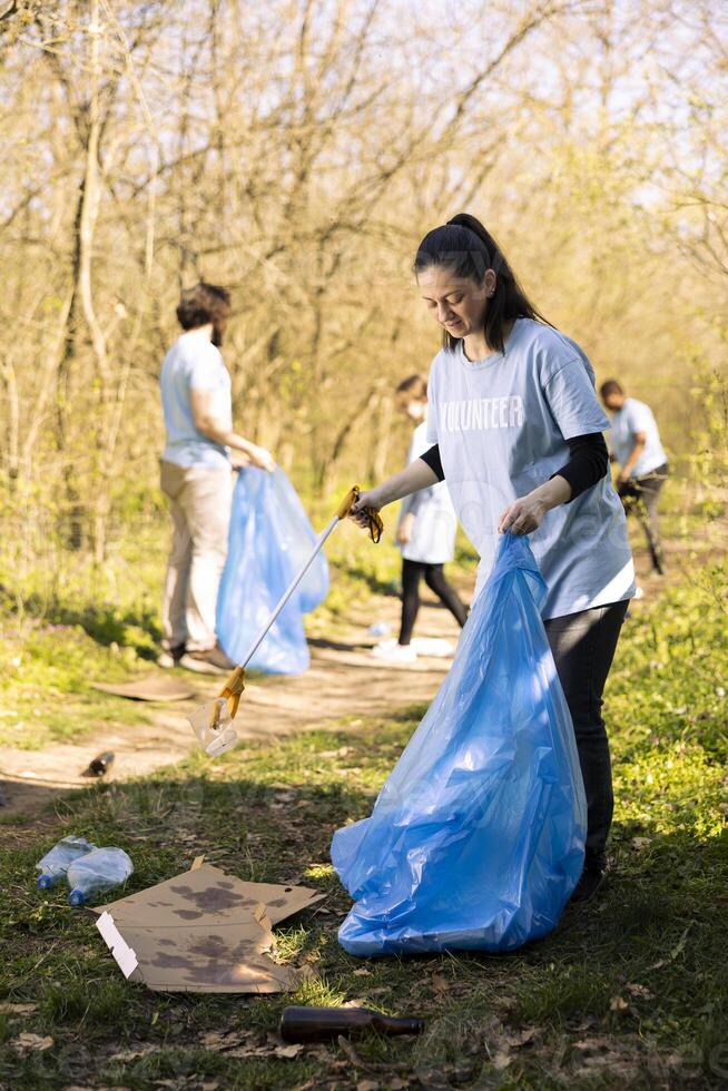 kvinna volontär- städa de skog av sopor och plast flaskor, samlar skräp med klo och förfogande väska. ung aktivist volontär till bevara naturlig ekosystem. foto