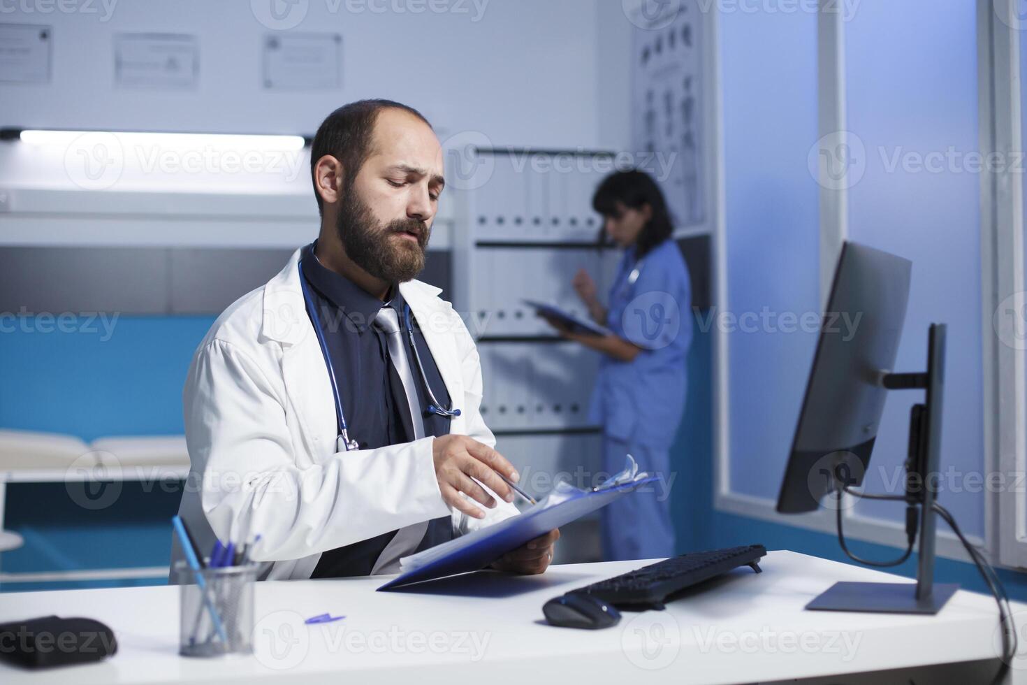 bild ställer ut en tillägnad caucasian läkare kontroll och analyserar hans anteckningar på de klinik kontor skrivbord. ung man bär en vit labb täcka framställning för medicinsk samråd med patienter. foto