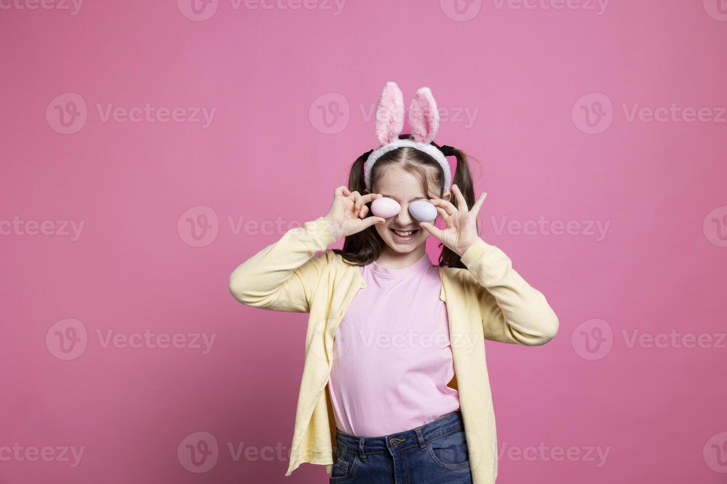 glad barn med kanin öron lurar runt om i studio, innehav handgjort målad påsk ägg till fira apirl fest. litet barn med pigtails Framställ med förtroende i främre av kamera. foto