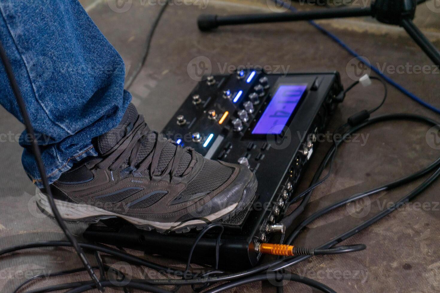 en fot steg på en gitarr effekter trampa, fångad från de sida. de sko är en enkel sneaker, och de trampa vilar på en texturerad golv. de fokus är på de handling, med en känsla av förväntan. foto