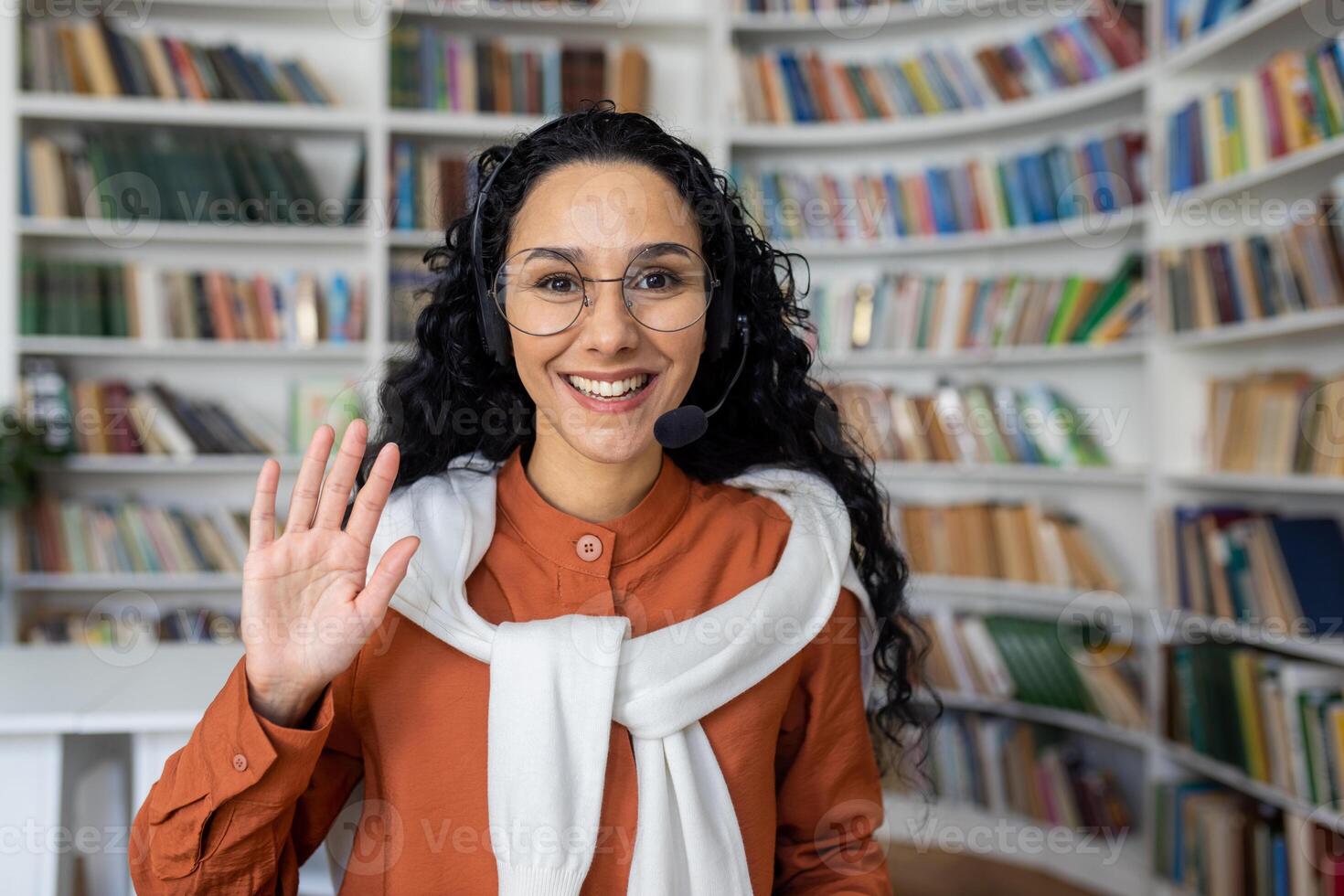 glad lockigt latinamerikan flicka Sammanträde på en tabell i främre av en bärbar dator i en headsetet ser på de kamera och vinka på de kamera, kontor arbetstagare i en ring upp Centrum, arbetssätt ring upp kamera se. foto