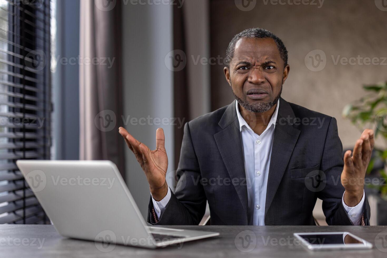förvirrad afrikansk amerikan affärsman i en kostym gester med osäkerhet på hans kontor skrivbord med bärbar dator och läsplatta. foto