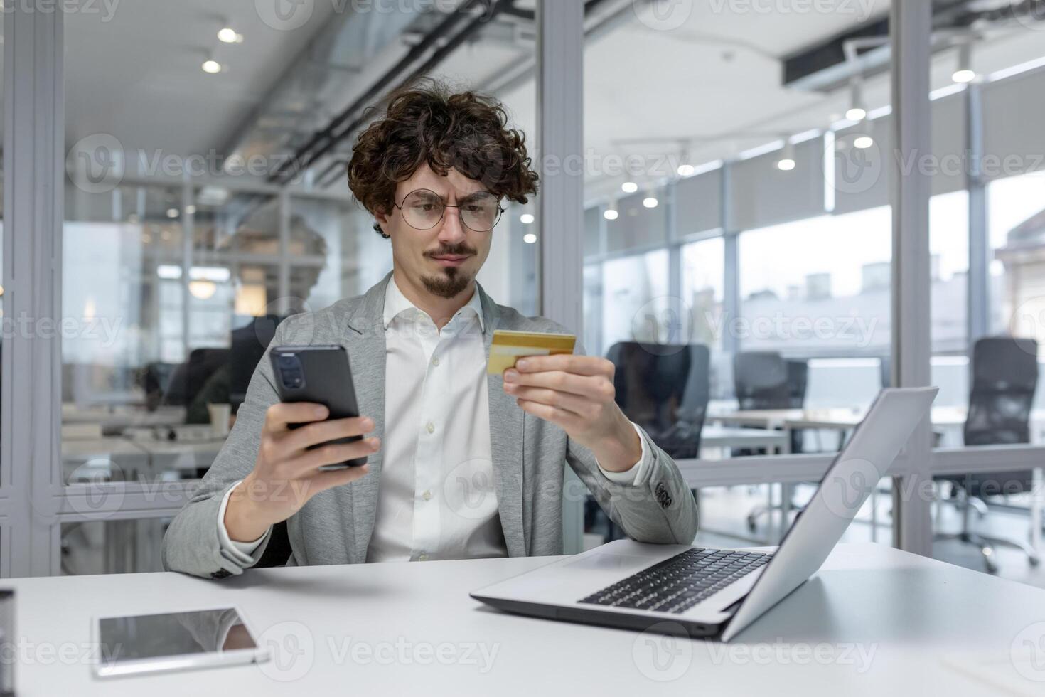 fokuserade ung affärsman med lockigt hår göra flera saker samtidigt i en modern kontor, arbetssätt på en bärbar dator medan kontroll hans telefon. foto