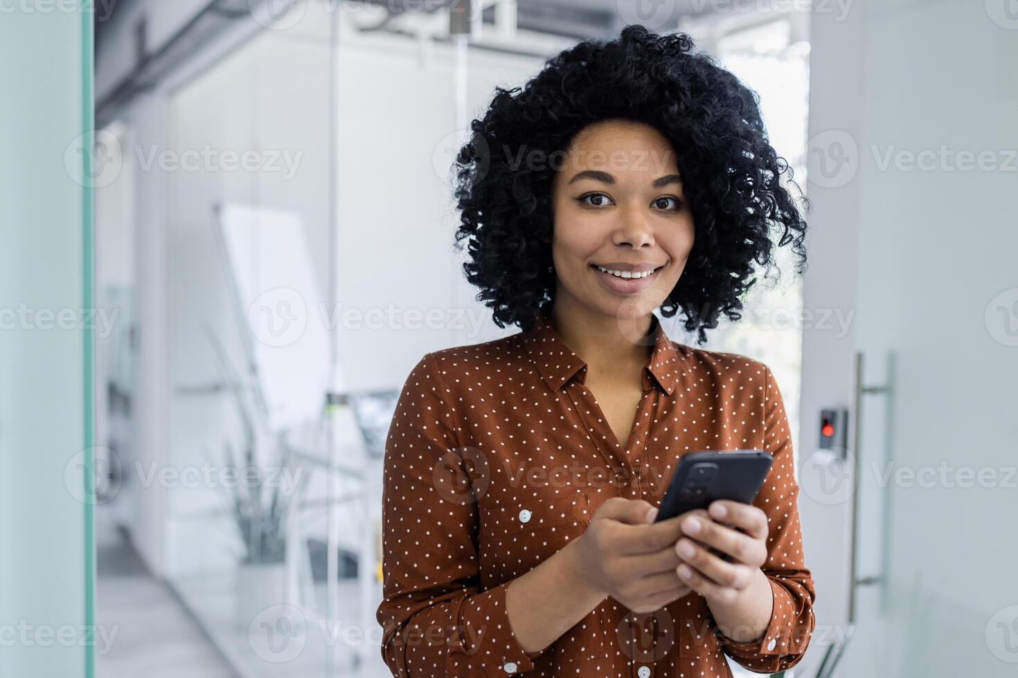 självsäker afrikansk amerikan affärskvinna använder sig av henne smartphone med en trevlig leende, stående i en ljus modern kontor. professionell klädsel och positiv arbetsplats energi. foto