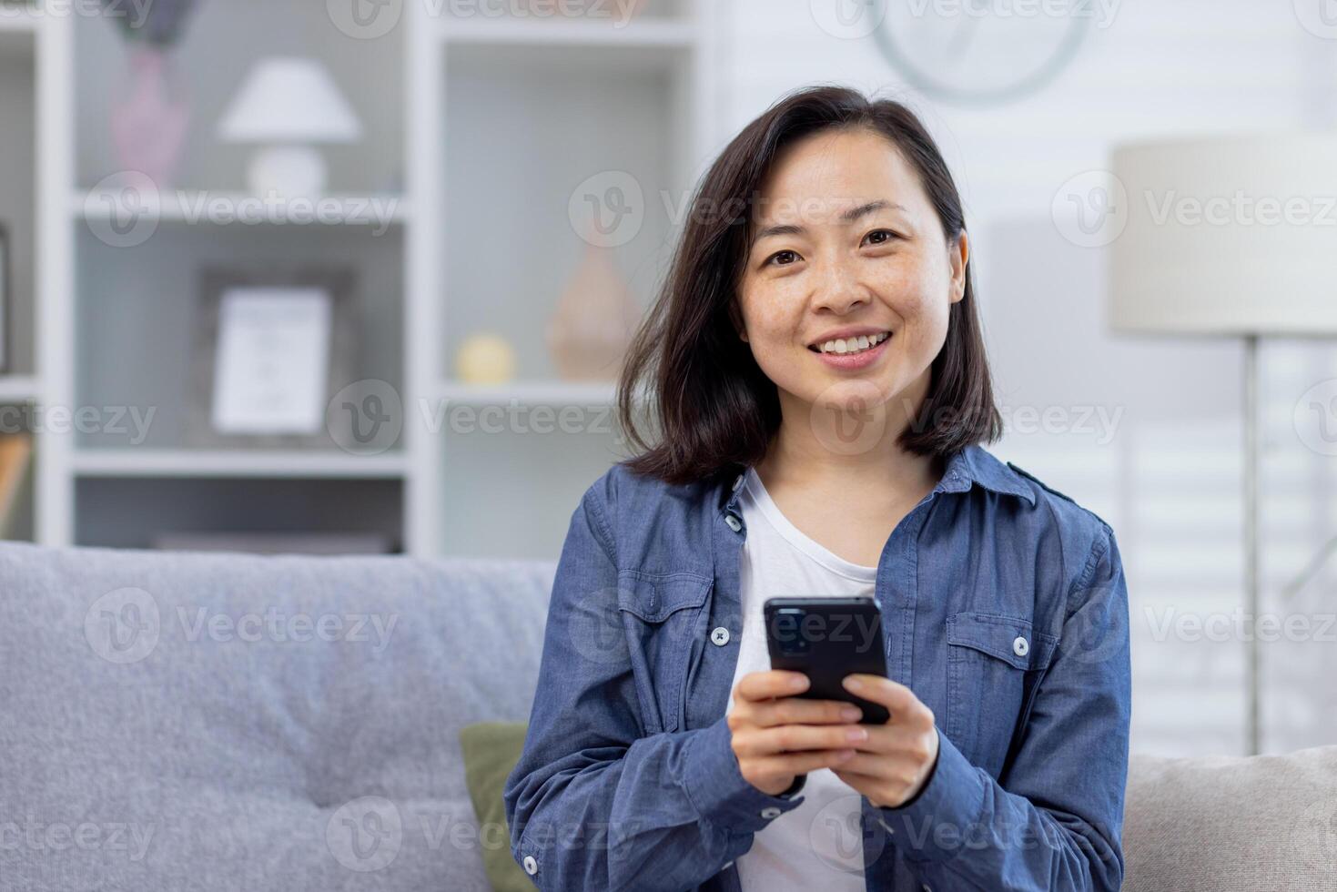 porträtt av en ung skön asiatisk kvinna på Hem på de soffa, kvinna leende och ser på de kamera innehav en telefon, smartphone Ansökan användare, nöjd och Lycklig. foto