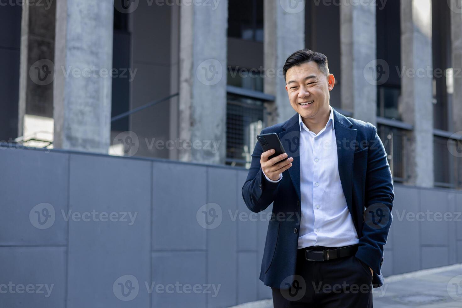 professionell asiatisk man i en eleganta kostym ler som han texter på hans telefon, stående på en solig urban trottoar. foto