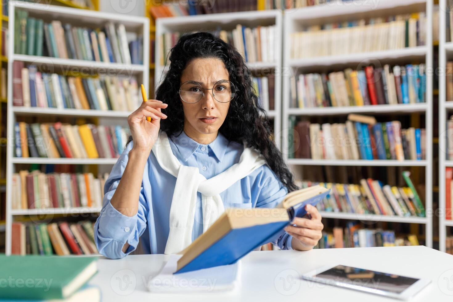 en allvarlig, medelålders kvinna bibliotekarie med glasögon försiktigt undersöker en bok medan organisering de bibliotek hyllor, omgiven förbi ett omfattande samling av böcker. foto