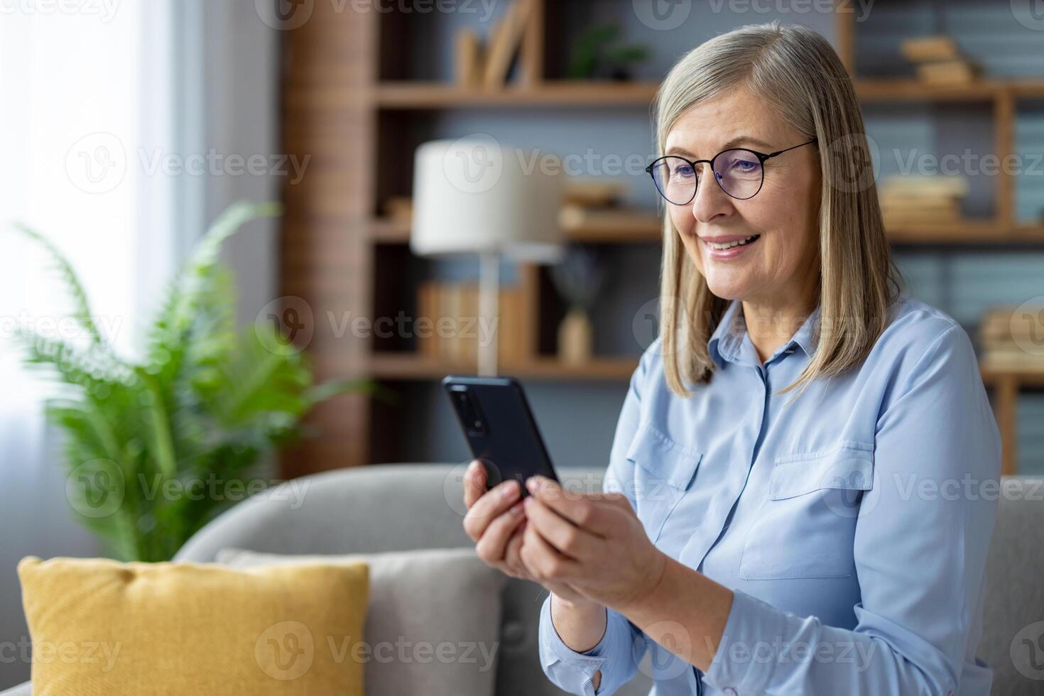 en medelålders kvinna i en blå skjorta Sammanträde på en soffa, absorberad i henne smartphone i en väl upplyst, mysigt levande rum miljö. foto