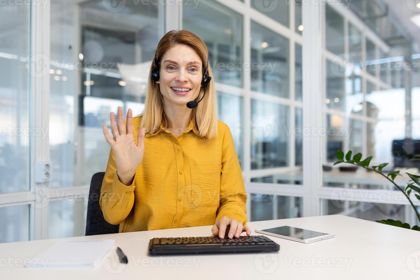 porträtt av en ung kvinna i en headsetet vem sitter i de kontor på de tangentbord och hälsar de kamera med henne hand. Arbetar i Stöd och bistånd service, telemarket, ring upp Centrum. foto