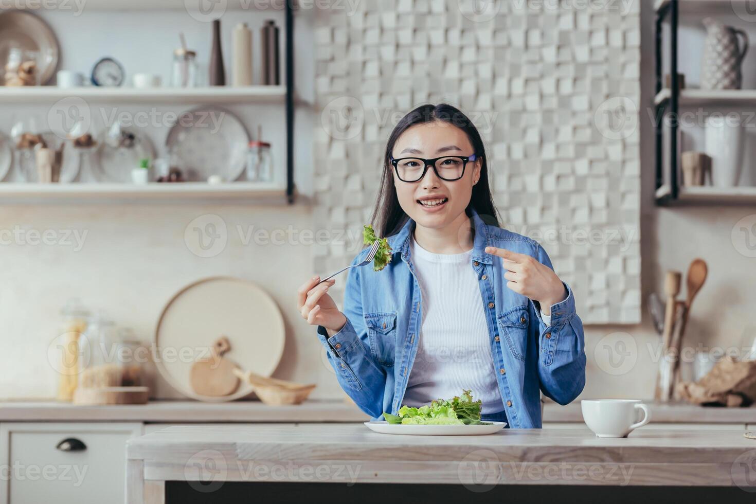 porträtt av Lycklig kvinna på Hem, asiatisk kvinna äter friska mat sallad och leende, hemmafru ser på kamera Sammanträde i kök på tabell, diet för vikt förlust och kondition. foto