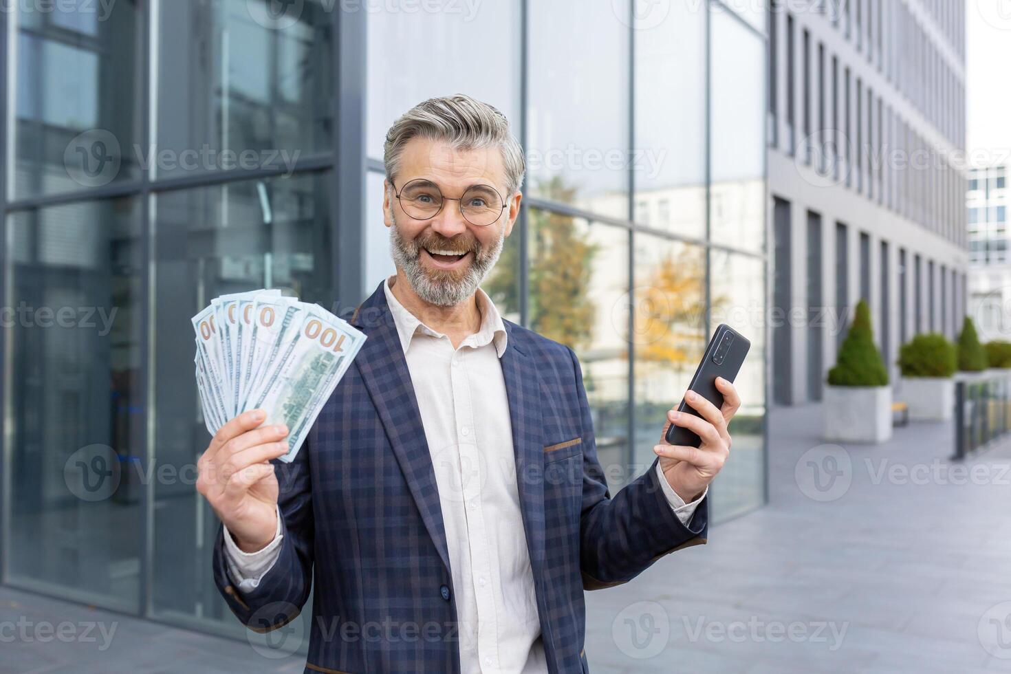 överraskad affärsman innehav lugg av pengar och telefon i händer, Lycklig med vinnande lotteri, befordran på arbete, ser på kamera och leende. foto