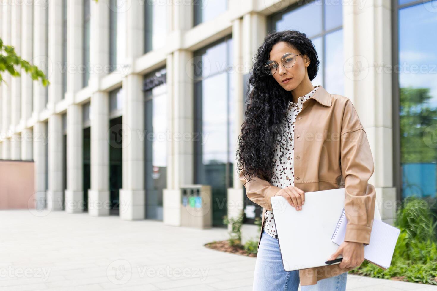 självsäker ung affärskvinna med bärbar dator och anteckningsbok stående i främre av ett kontor byggnad, utsöndrar professionalism och ambition. foto