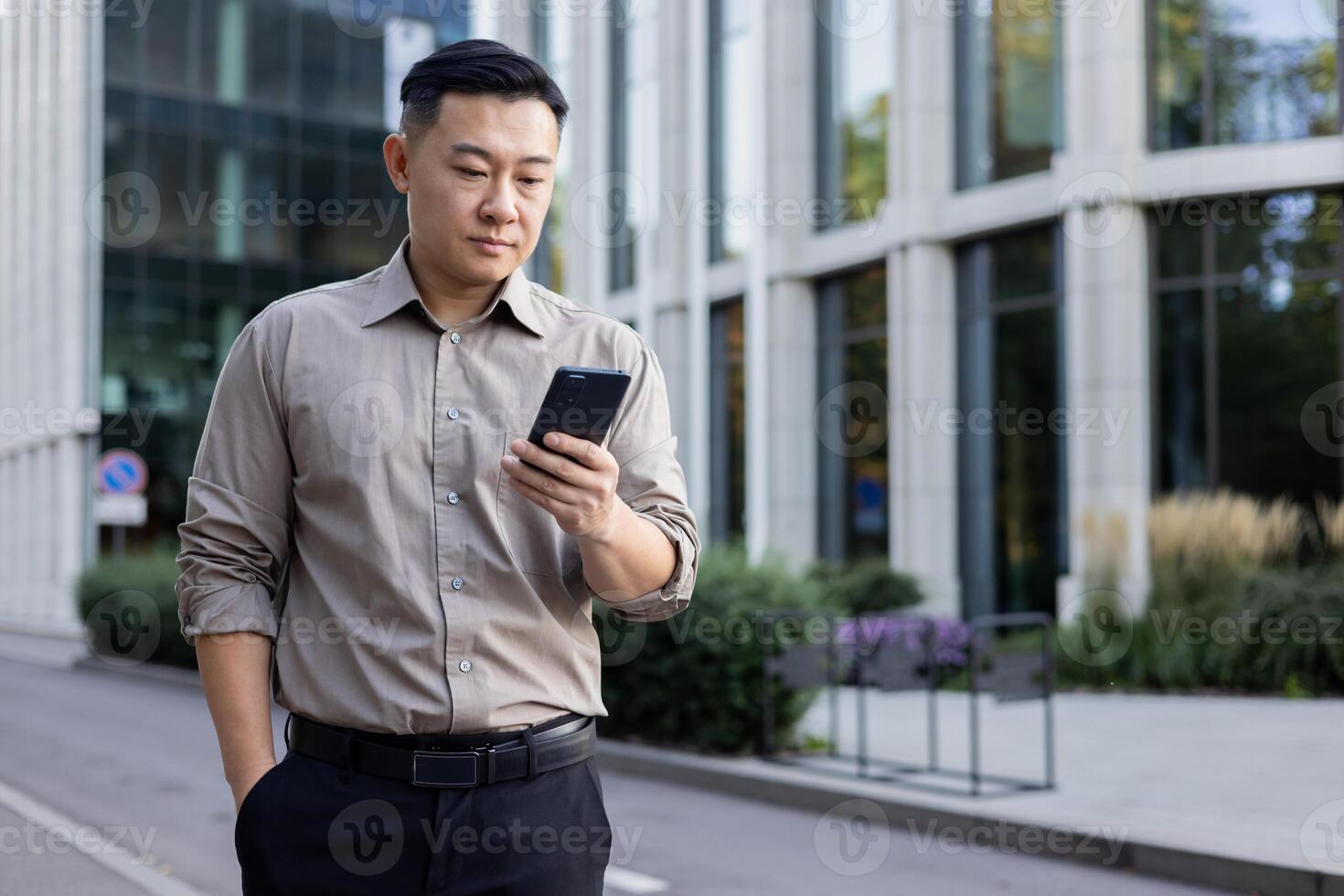 allvarlig ung asiatisk manlig advokat stående nära kontor byggnad och använder sig av mobil telefon. foto