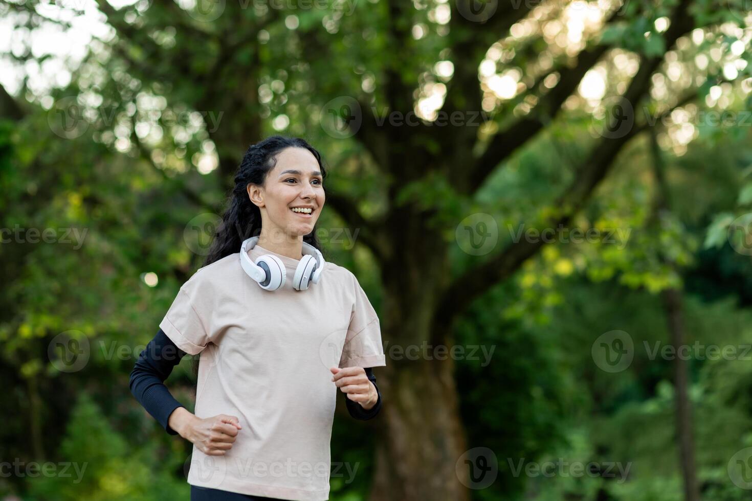 aktiva kvinna i t-shirt över lång ärm med headsetet över nacke utövar utomhus under dagtid i grön område. positiv lady håller på med joggning i offentlig parkera för förvaring passa och friska kropp. foto