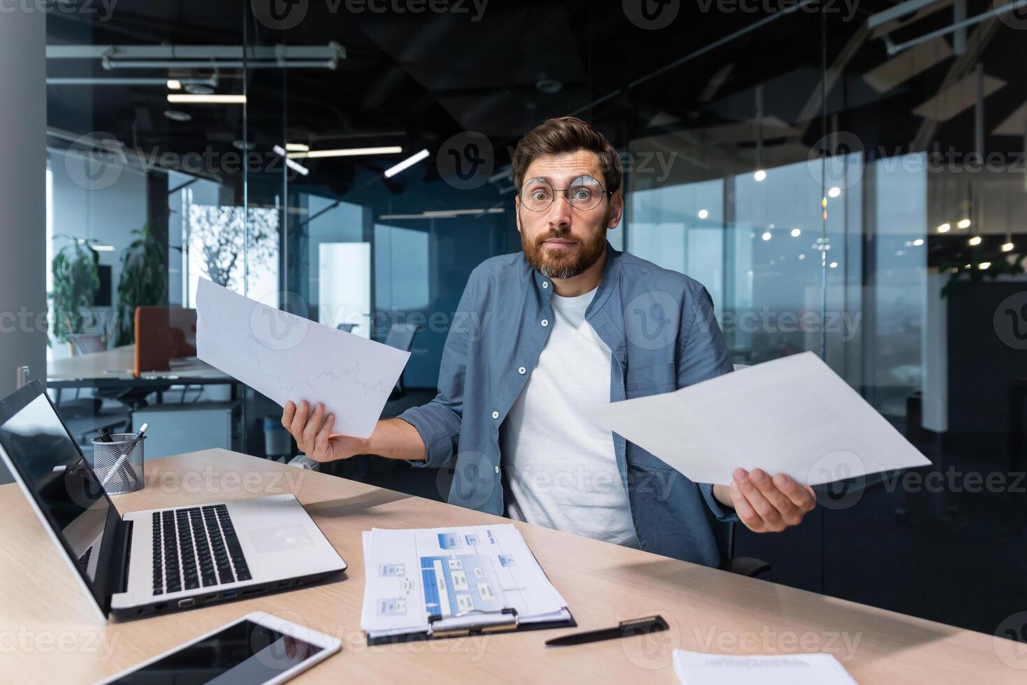 missnöjd och arg affärsman inuti kontor som visar rapporter och räkningar dokument till kamera, chef i skjorta ser på kamera arbetssätt med bärbar dator papper arbete. foto