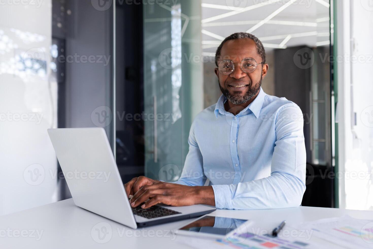 porträtt av mogna affärsman inuti modern kontor, senior man i skjorta leende och ser på kamera, arbetstagare använder sig av bärbar dator på arbete foto