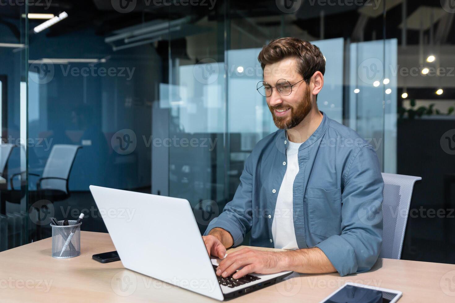 leende och glad affärsman i tillfällig skjorta arbetssätt inuti modern kontor, mogna programmerare skrivning koda använder sig av bärbar dator. foto