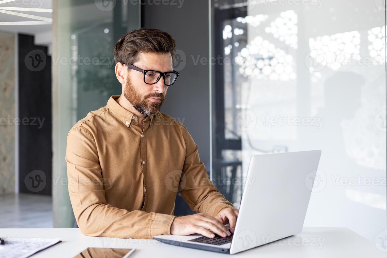 allvarlig koncentrerad tänkande affärsman arbetssätt inuti kontor med bärbar dator, mogna upplyst man i skjorta och glasögon på arbetsplats skriver på tangentbord, programmerare utvecklaren. foto