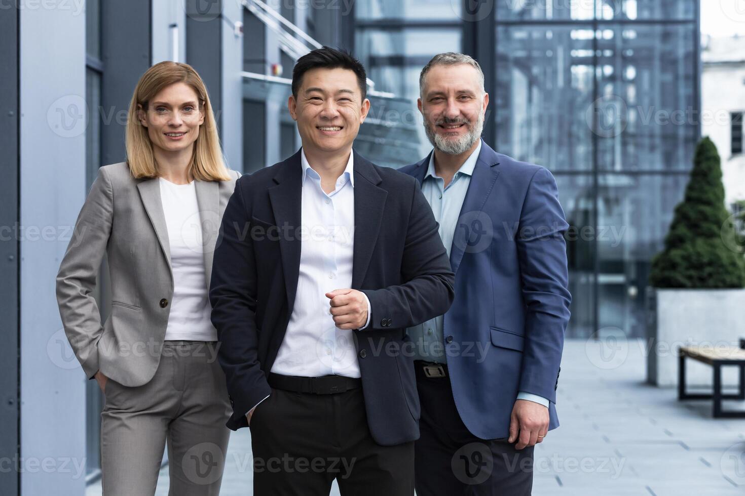 framgångsrik olika företag team, tre arbetare leende och ser på kamera, dröm team med asiatisk chef utanför kontor byggnad, kollegor i företag kostymer foto