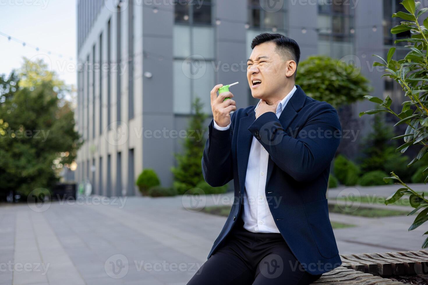 lidande från en öm hals, en ung asiatisk manlig affärsman sitter på en bänk utanför ett kontor Centrum och sprayer medicin aerosol ner hans hals, innehav hans hand till hans nacke. foto