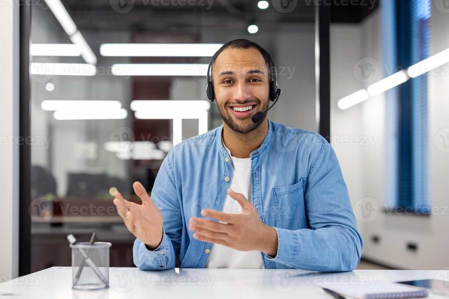 porträtt av en leende ung latinamerikan man bär en headsetet Sammanträde på ett kontor skrivbord och talande till de kamera. foto