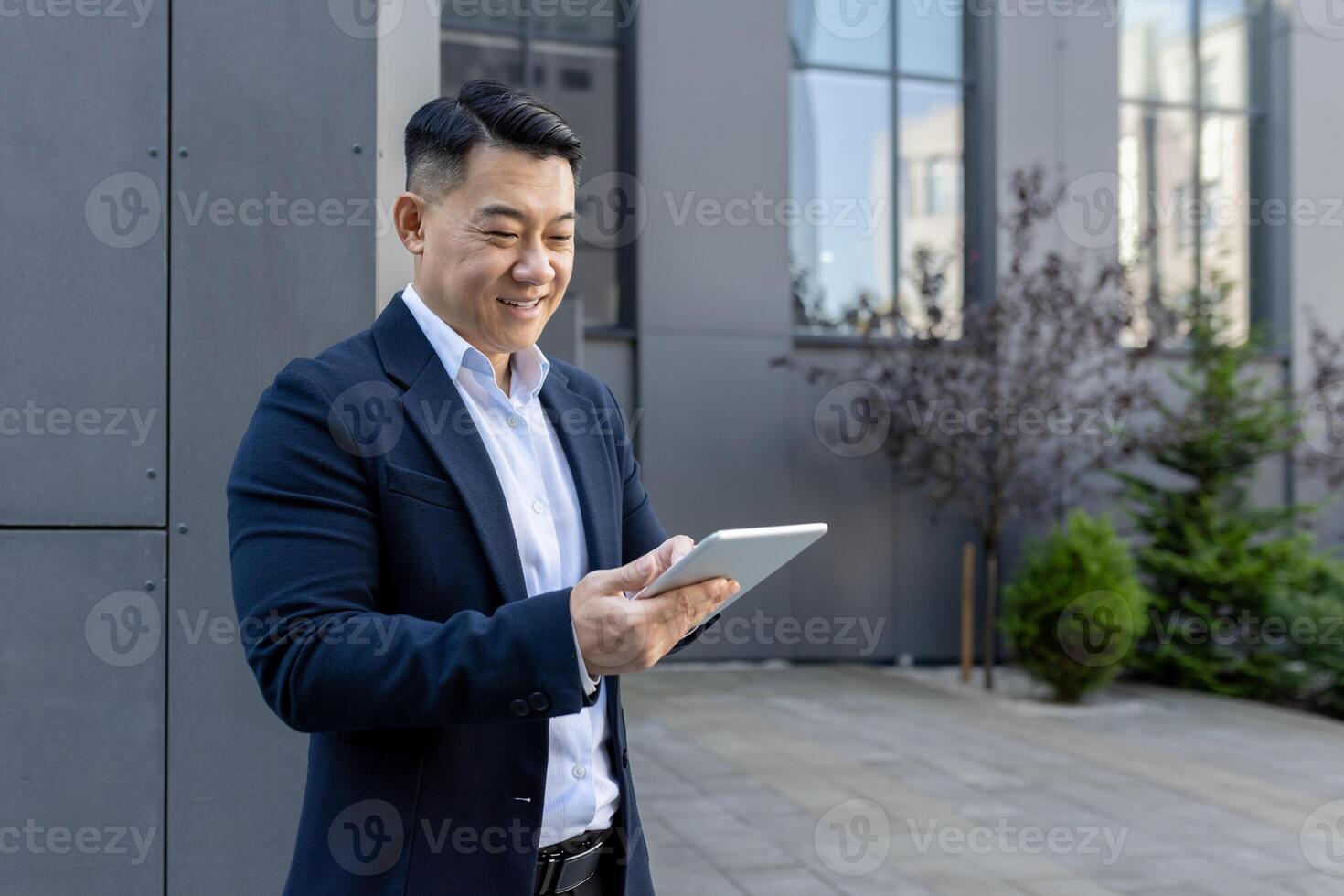 leende asiatisk manlig professionell i en kostym engagerande med en läsplatta pc på en solig dag utanför en företags- byggnad. foto