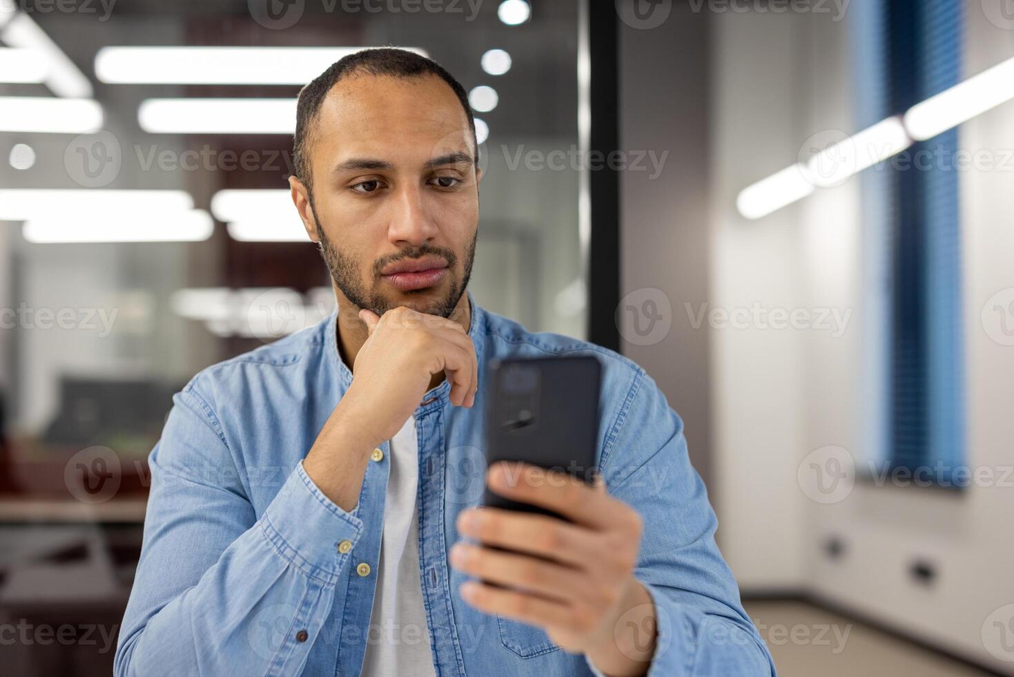 en vardagligt klädd man engagerar med hans smartphone, bläddring eller kommunicera, medan stående i en samtida kontor miljö med en självsäker hållning. foto