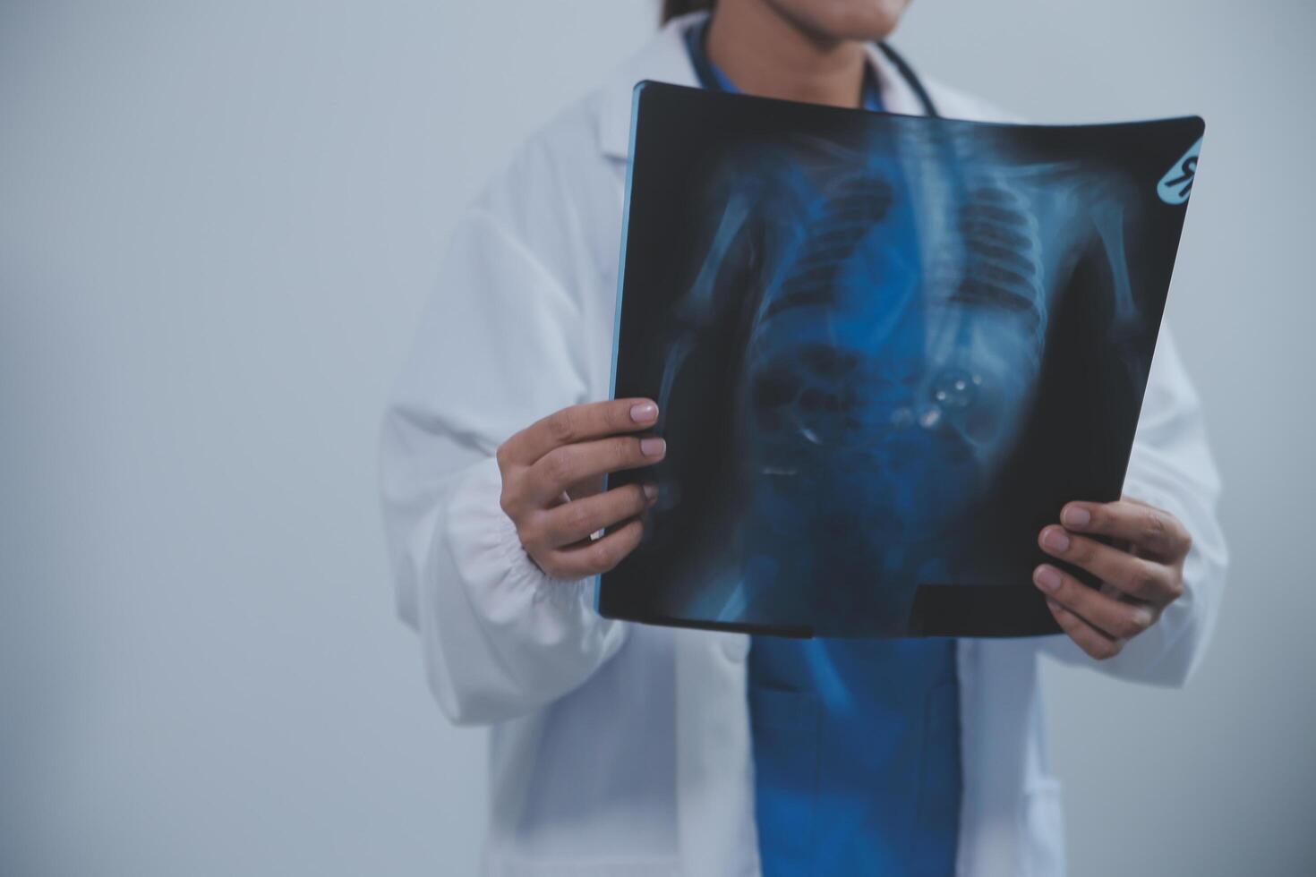 asiatisk muslim läkare i hijab och scrubs hörlurar runt om henne nacke stå självsäkert i de medicinsk kontor, isolerat på vit bakgrund, innehav röntgen filma. foto