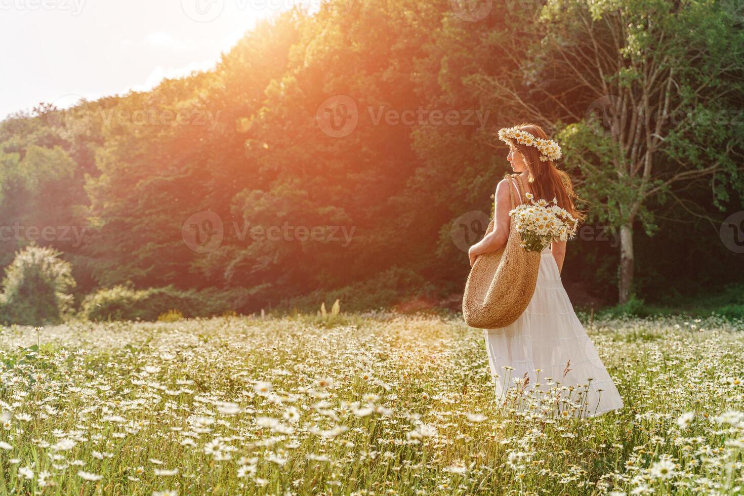 en ung kvinna i en vit solklänning, en krans av daisy med en stor korg- väska på henne axel är gående genom en fält av prästkragar, mot de bakgrund av de skog, de solnedgång ljus foto