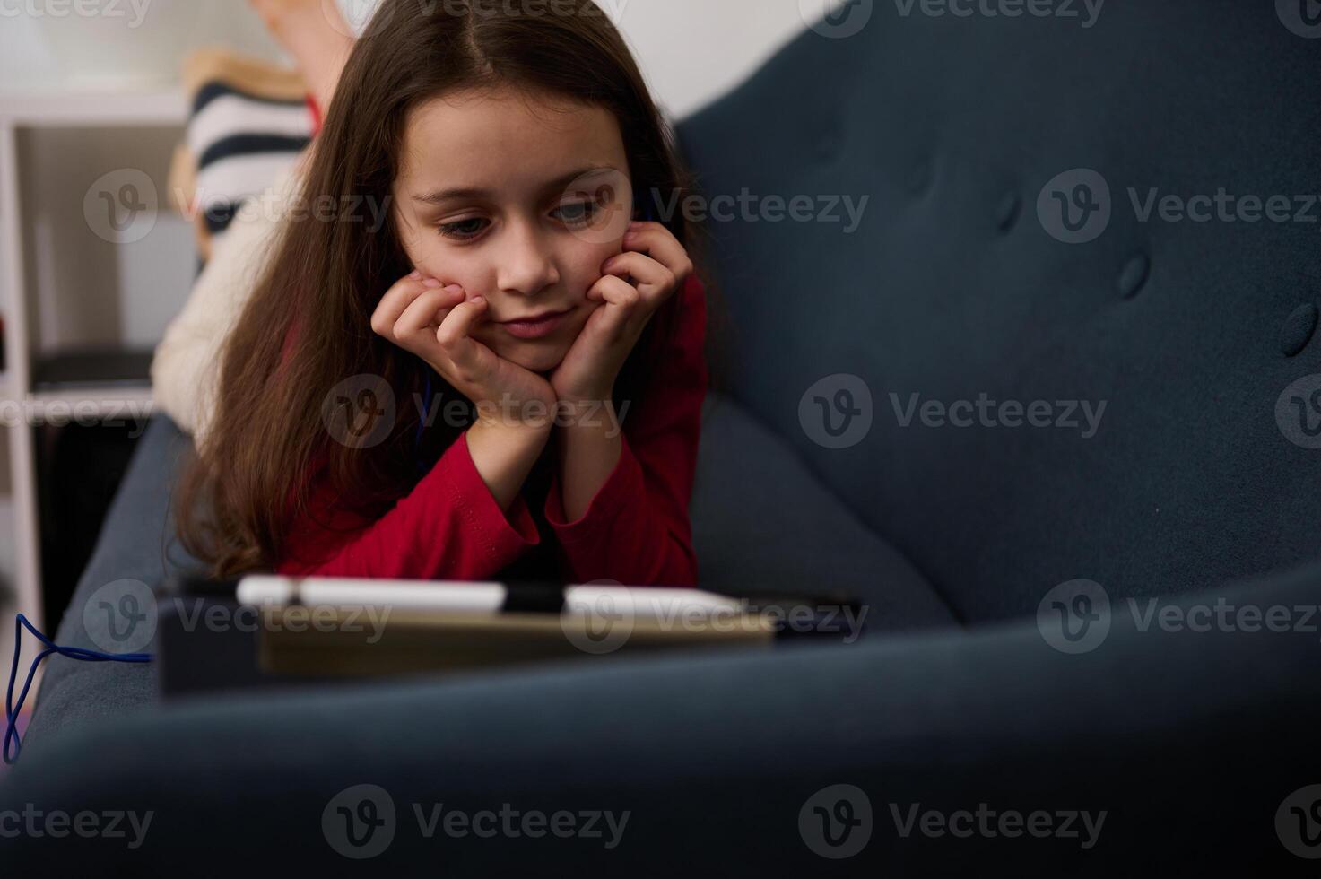 närbild caucasian elementärt ålder skolflicka, en härlig liten unge flicka har roligt på henne smart digital läsplatta, liggande på de soffa i henne helgen. liten barn tittar på tecknad serie på digital grej foto