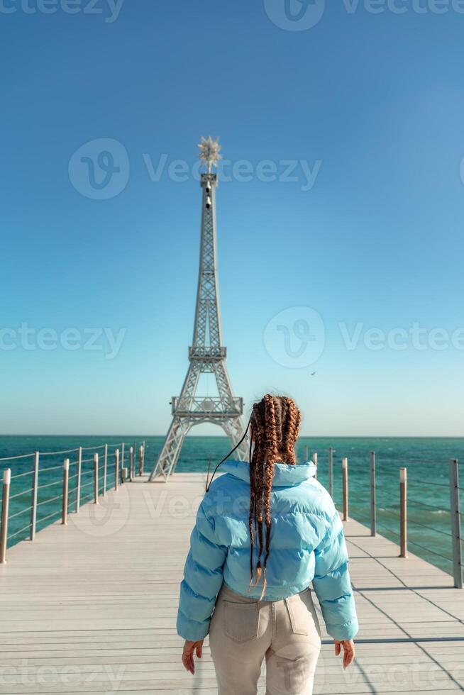 stor modell av de eiffel torn på de strand. en kvinna promenader längs de pir mot de torn, bär en blå jacka och vit jeans. foto