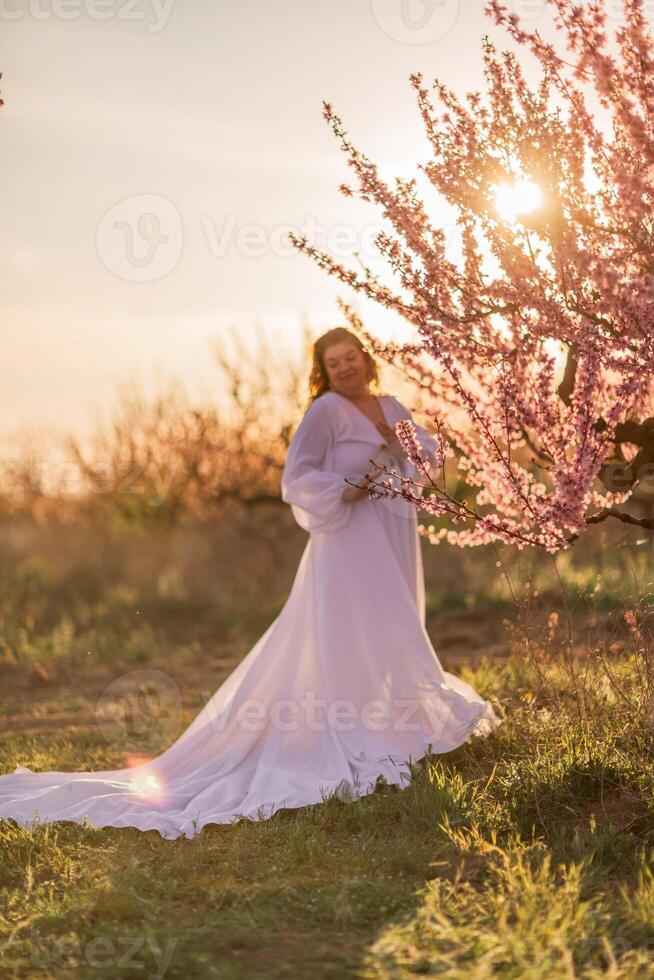 kvinna blomning persika fruktträdgård. mot de bakgrund av en pittoresk persika fruktträdgård, en kvinna i en lång vit klänning åtnjuter en fredlig promenad i de parkera, omgiven förbi de skönhet av natur. foto