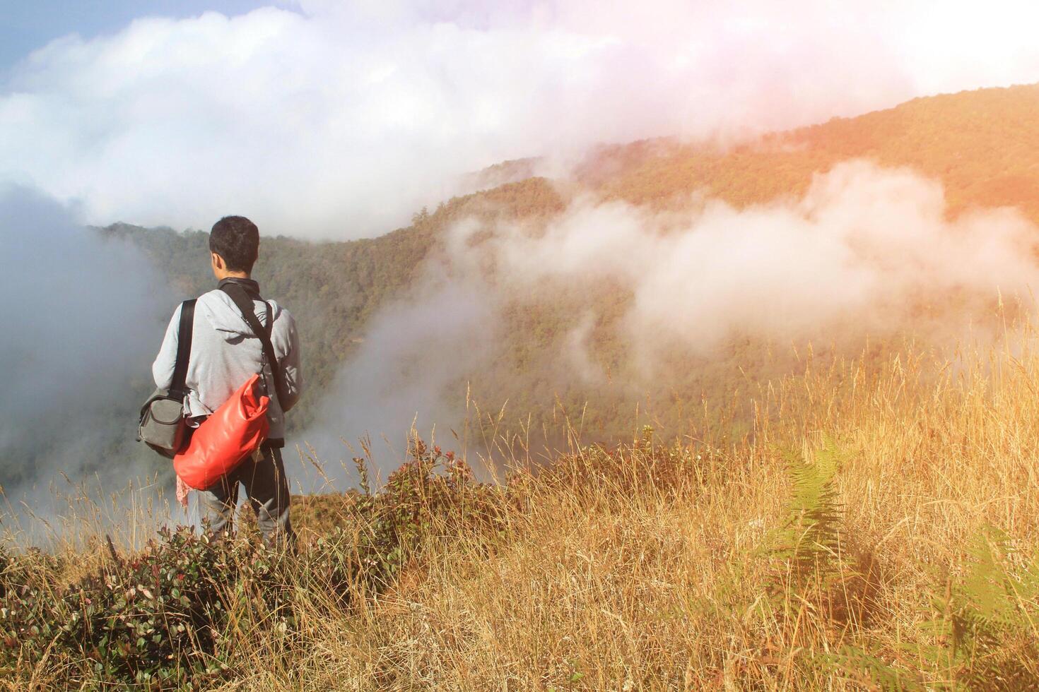 asiatisk man stående, bärande röd väska och ser till trevlig se, blå himmel, moln och dimma på berg synpunkt. resande och backpacker i natur med kopia Plats. foto