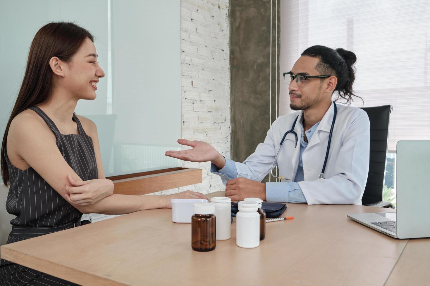 medicinsk behandling och kontroll, ung manlig läkare pratar med ett leende och undersöker en kvinnlig patient med asiatisk etnicitet under ett hälsokonsultationsbesök, råd på en sjukhusklinik. foto