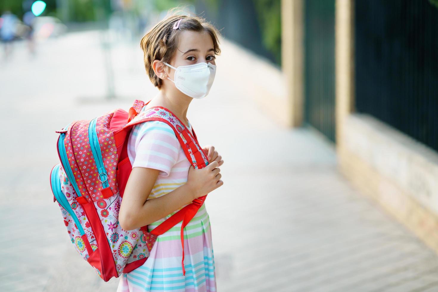 nio år gammal flicka går tillbaka till skolan iklädd en mask och en skolväska. foto
