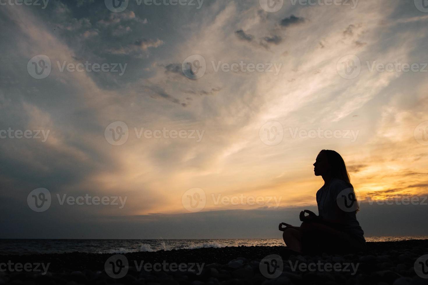 siluett av en kvinna i meditation poserar på havsstranden under surrealistisk solnedgång på havsbakgrund och dramatisk himmel foto