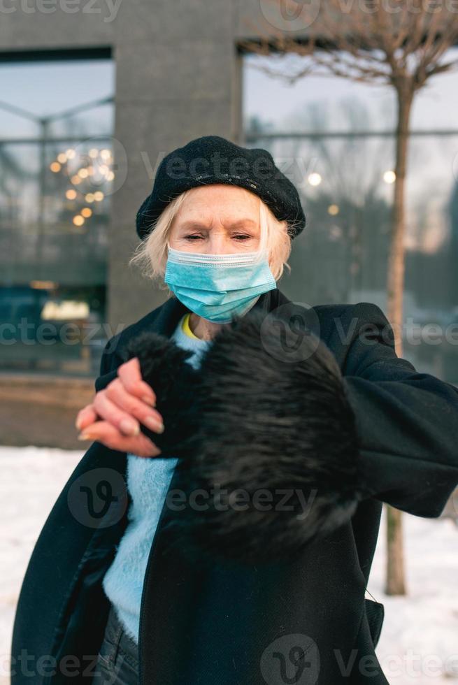 senior elegant kvinna i basker och elegant svart kappa och i medicinsk mask som går utomhus. lockdown, pandemi, skyddskoncept foto