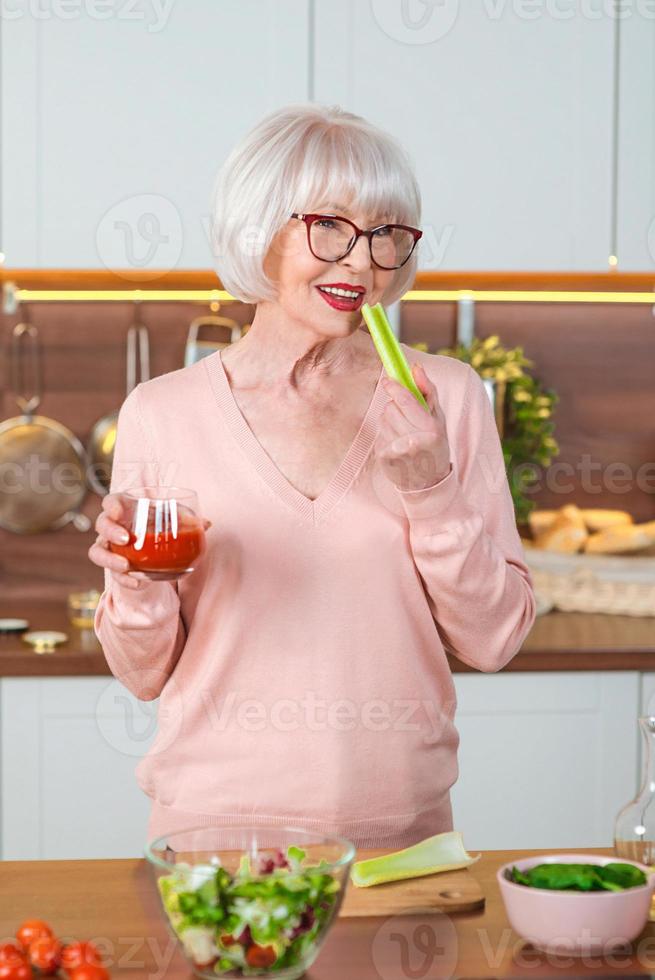 senior glad kvinna med selleri och tomatjuice på diet i hennes kök. rå, vegetarisk, kost, kökskoncept foto