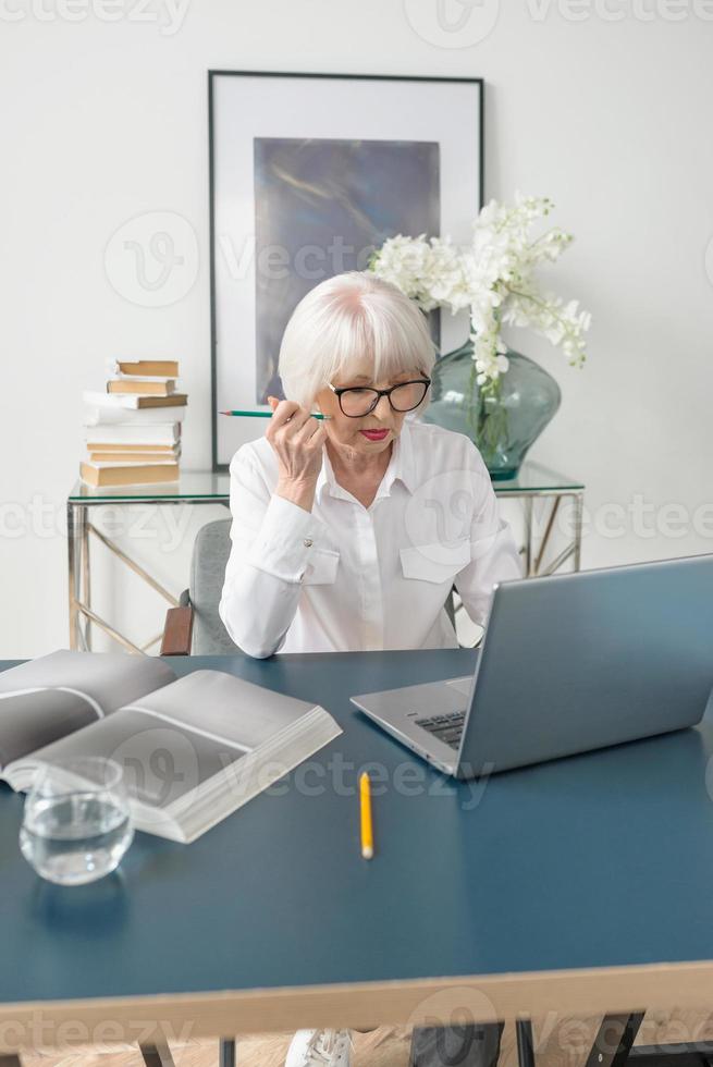 trött senior vacker grått hår kvinna i vit blus läser dokument på kontoret. arbete, seniorer, frågor, hitta en lösning, uppleva koncept foto