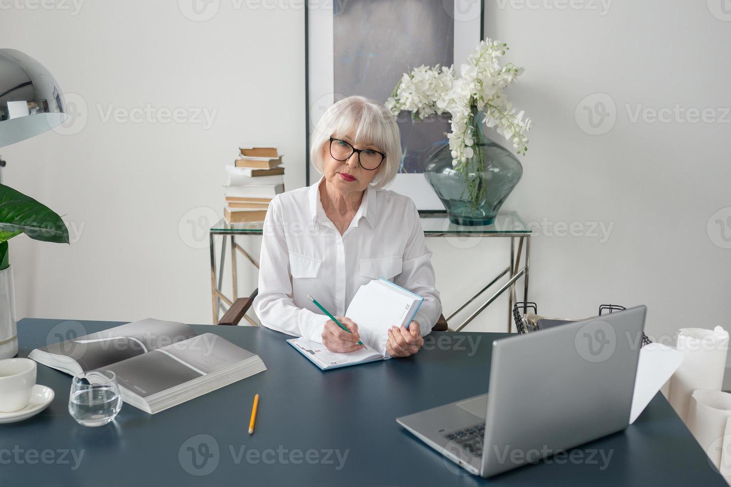 trött senior vacker grått hår kvinna i vit blus läser dokument på kontoret. arbete, seniorer, frågor, hitta en lösning, uppleva koncept foto