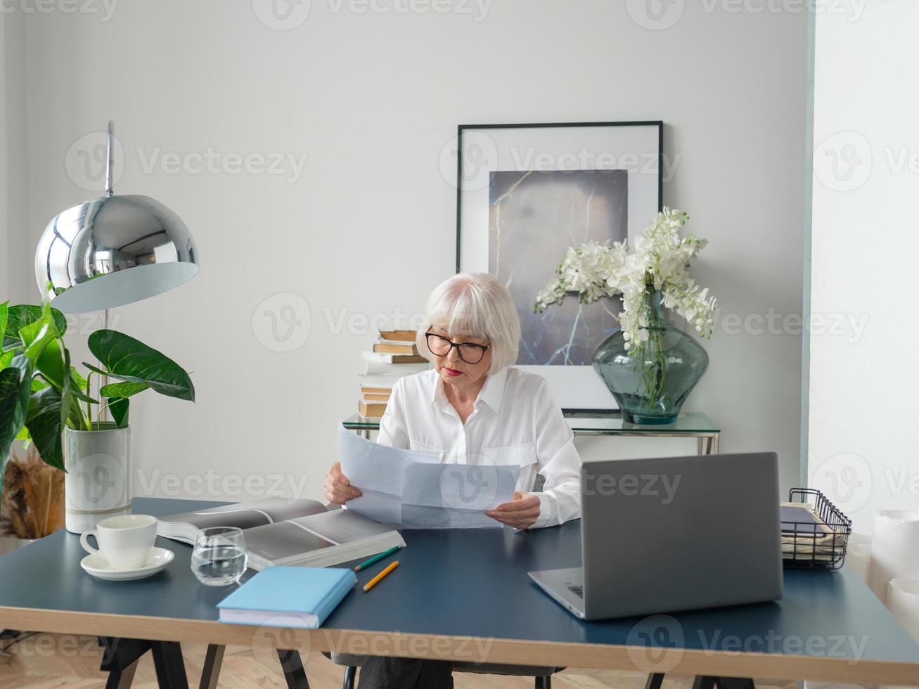 trött senior vacker grått hår kvinna i vit blus läser dokument på kontoret. arbete, seniorer, frågor, hitta en lösning, uppleva koncept foto